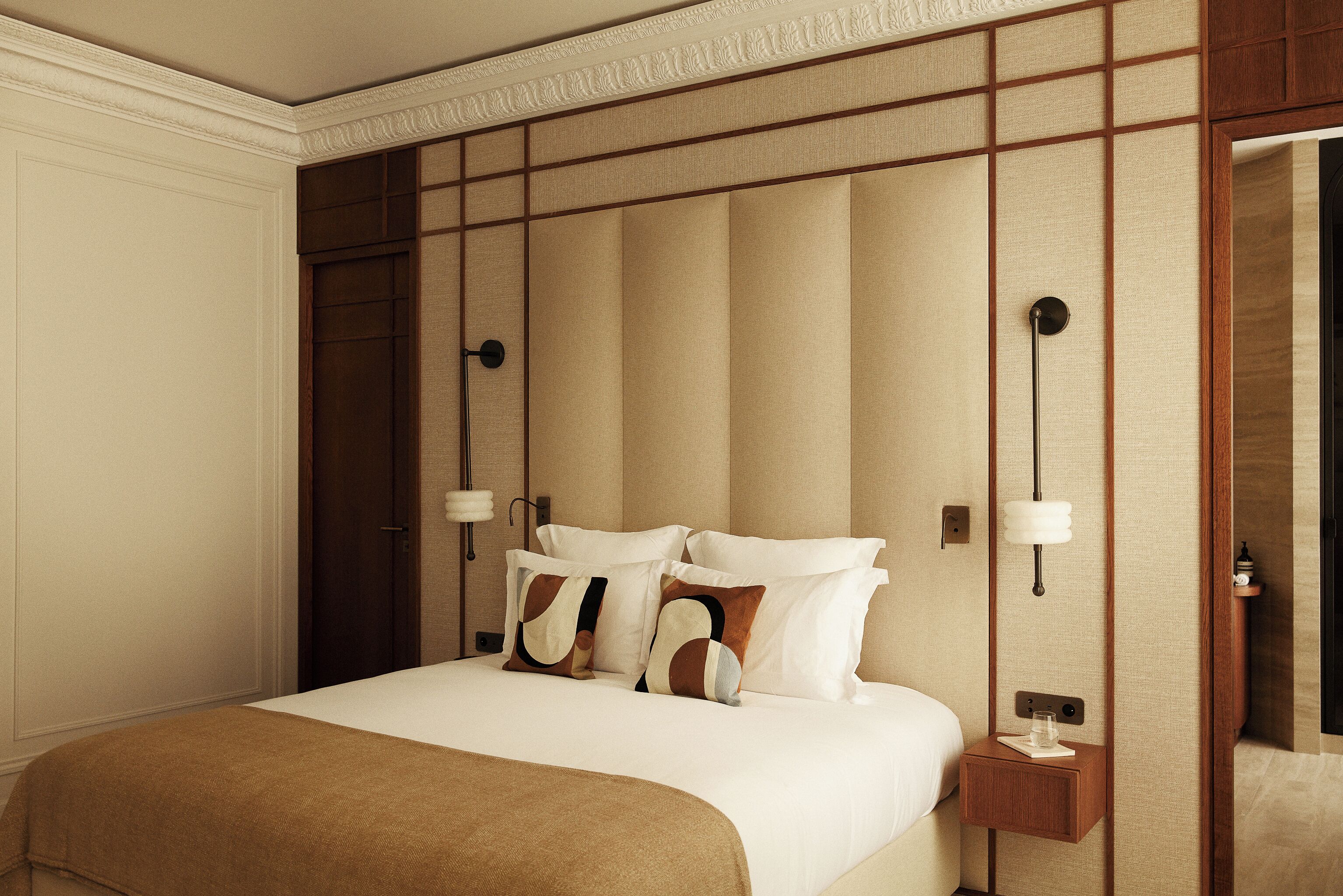 A neatly made bed with a brown throw blanket, a wooden nightstand with a lamp, and a mirror on the wall.
