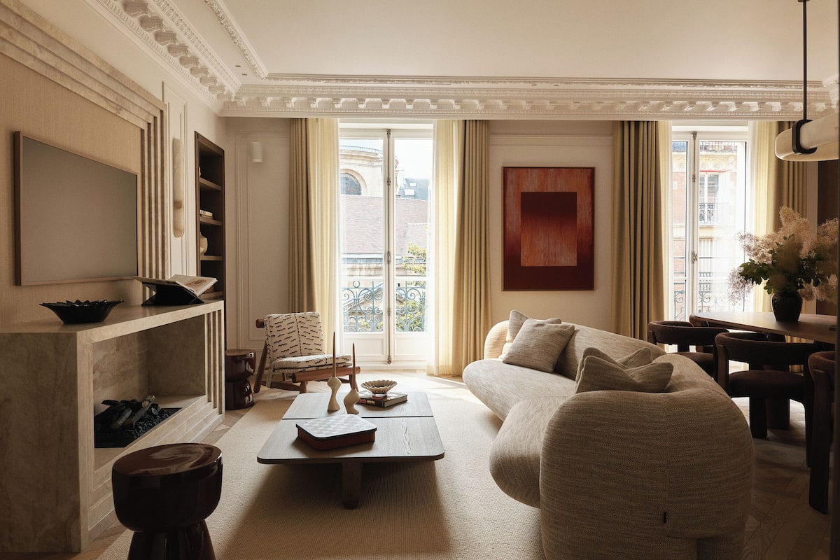 A spacious living room with a large sofa, a coffee table, and a television. There is a balcony with a view of the city.