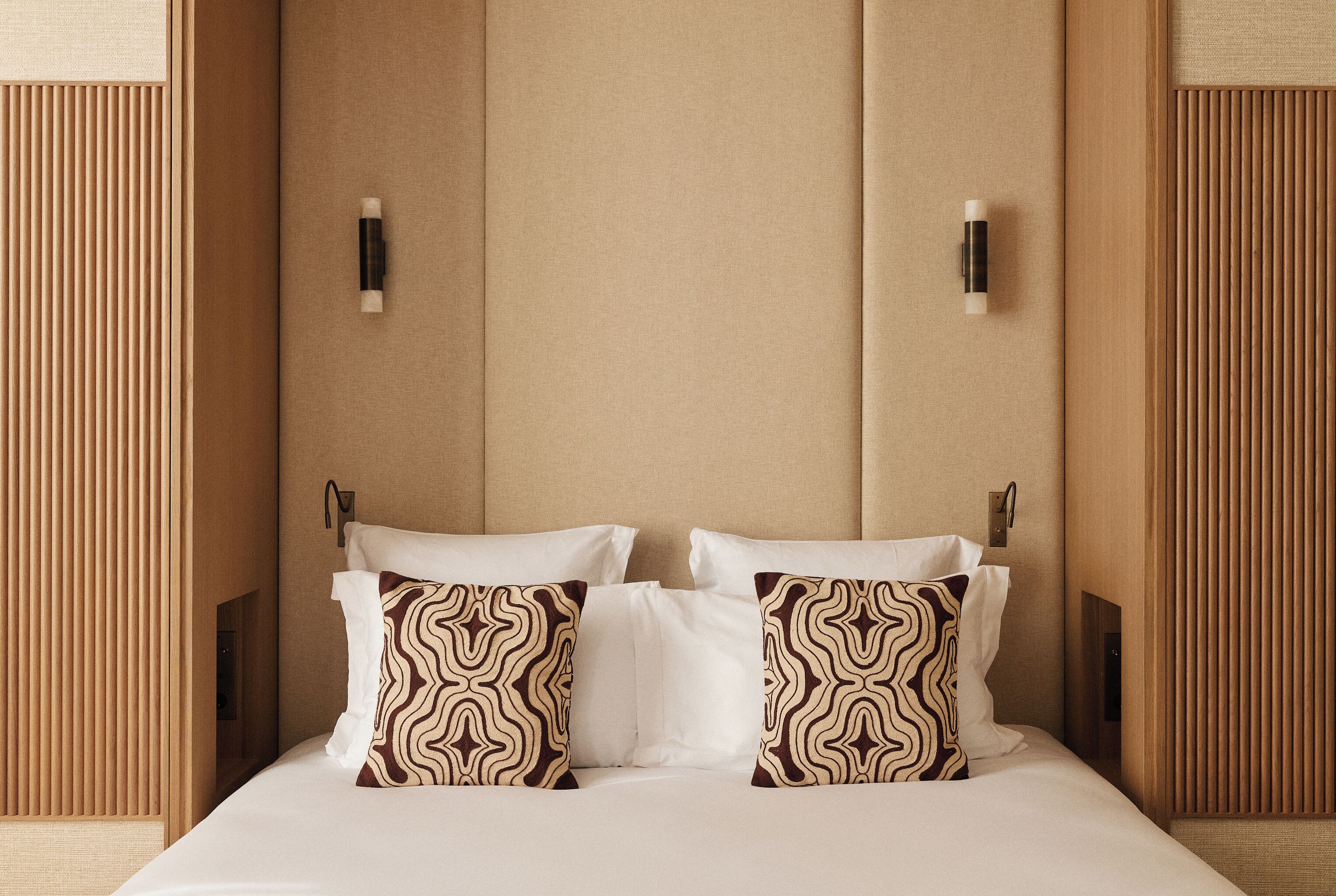 Une chambre d’hôtel avec un grand lit, deux coussins à motifs, une tête de lit à lattes verticales et des appliques murales.