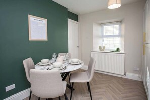 Un comedor con una mesa dispuesta para cuatro, sillas, un cuadro en la pared, una ventana con persianas y un radiador.