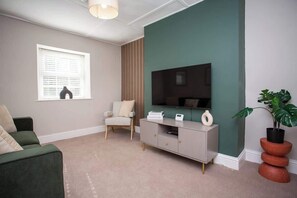 Una sala de estar con una pared de acento verde, un televisor de pantalla plana, un mueble gris para entretenimiento, un sofá verde, un sillón blanco y una planta en maceta.