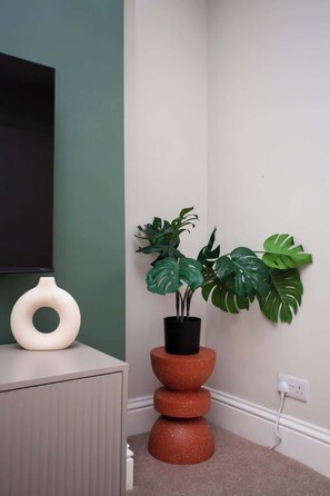 Un rincón con una pared verde, un jarrón blanco, una planta en maceta y un enchufe.