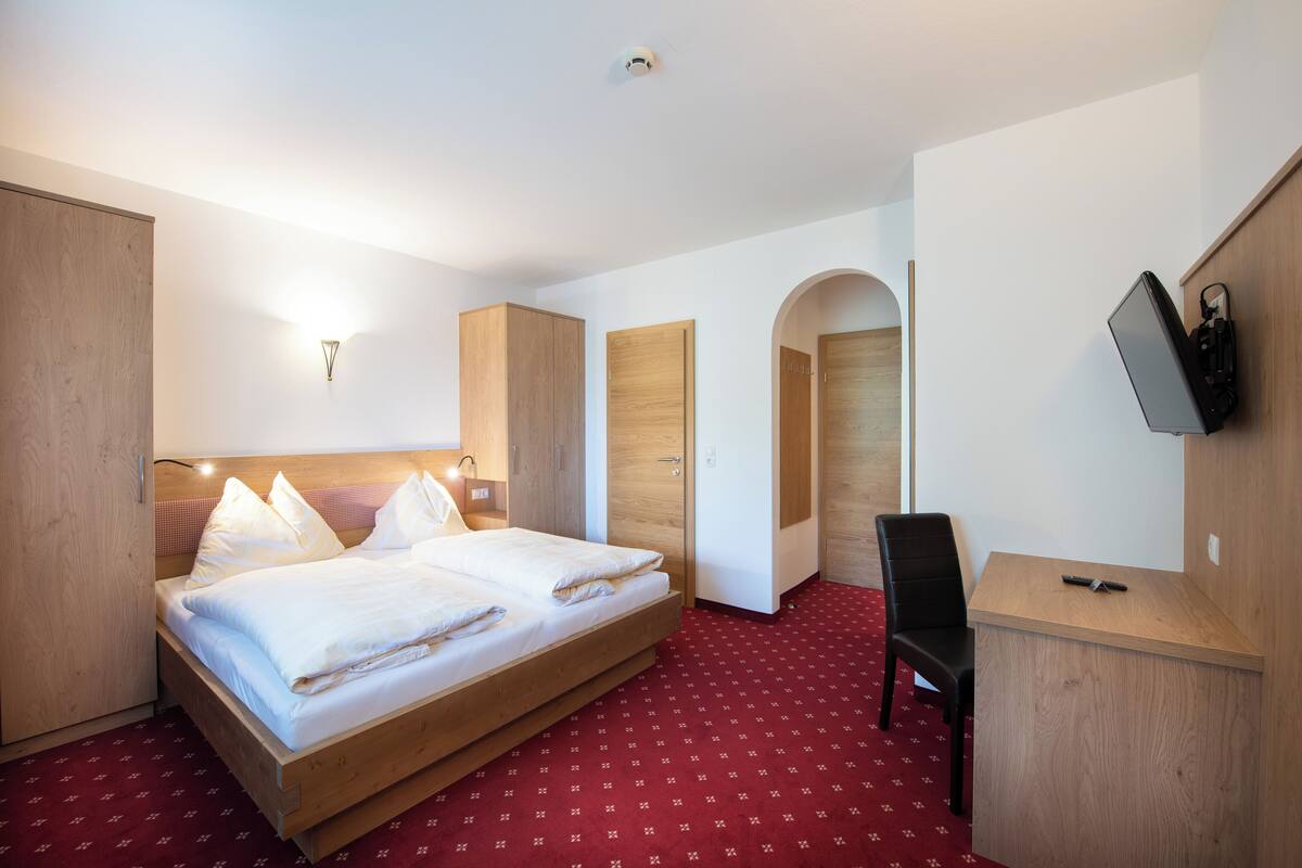 A hotel room with a wooden bed, a desk with a chair, a TV mounted on the wall, and a red carpet.