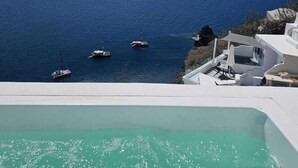 A clear view of a turquoise sea with boats, a white infinity pool, and a white building with a balcony overlooking the sea.
