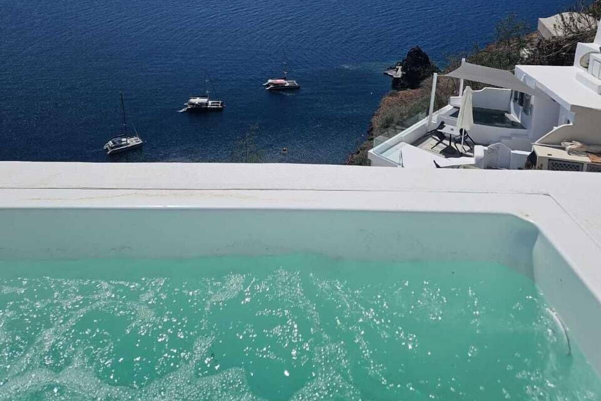 Una vista chiara su un mare turchese con barche, una piscina a sfioro bianca e un edificio bianco con un balcone che si affaccia sul mare.