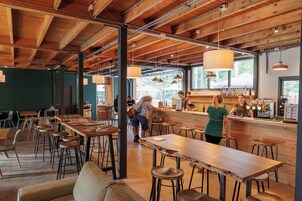 Um café amplo com mesas e cadeiras de madeira, um espaço de bar e janelas grandes que permitem entrada de luz natural.