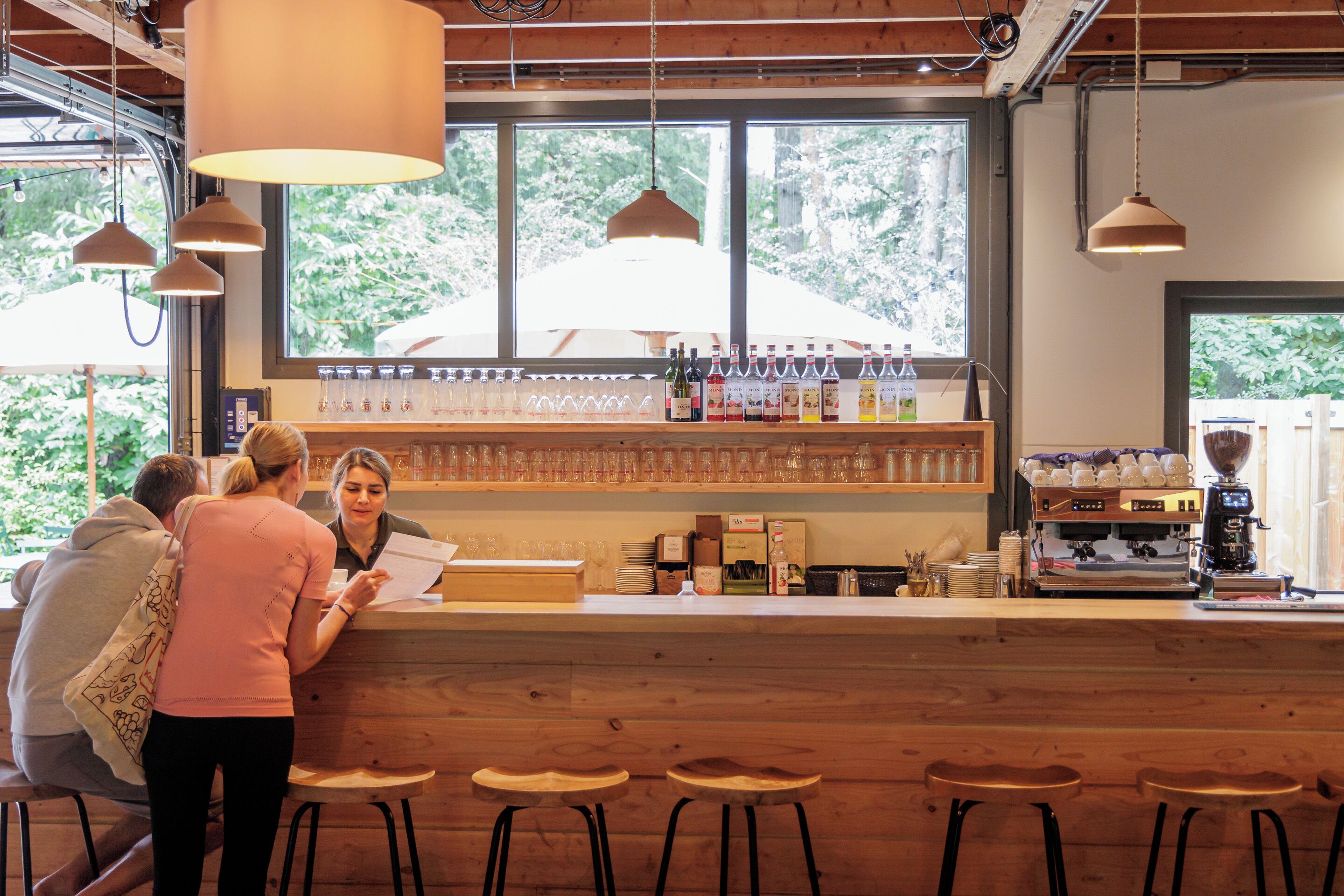 Eine Bar mit Holzhockern, einer Theke mit Kaffeemaschine und Regalen voller Flaschen und Gläser.
