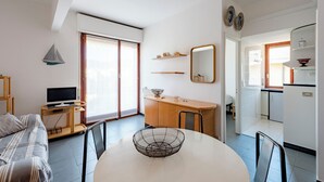 Ein modernes, helles Wohnzimmer mit einer Couch, einem Esstisch mit einer dekorativen Schale, einem Fernseher auf einem Schrank und einem durch eine offene Tür einsehbarer Küchenbereich.