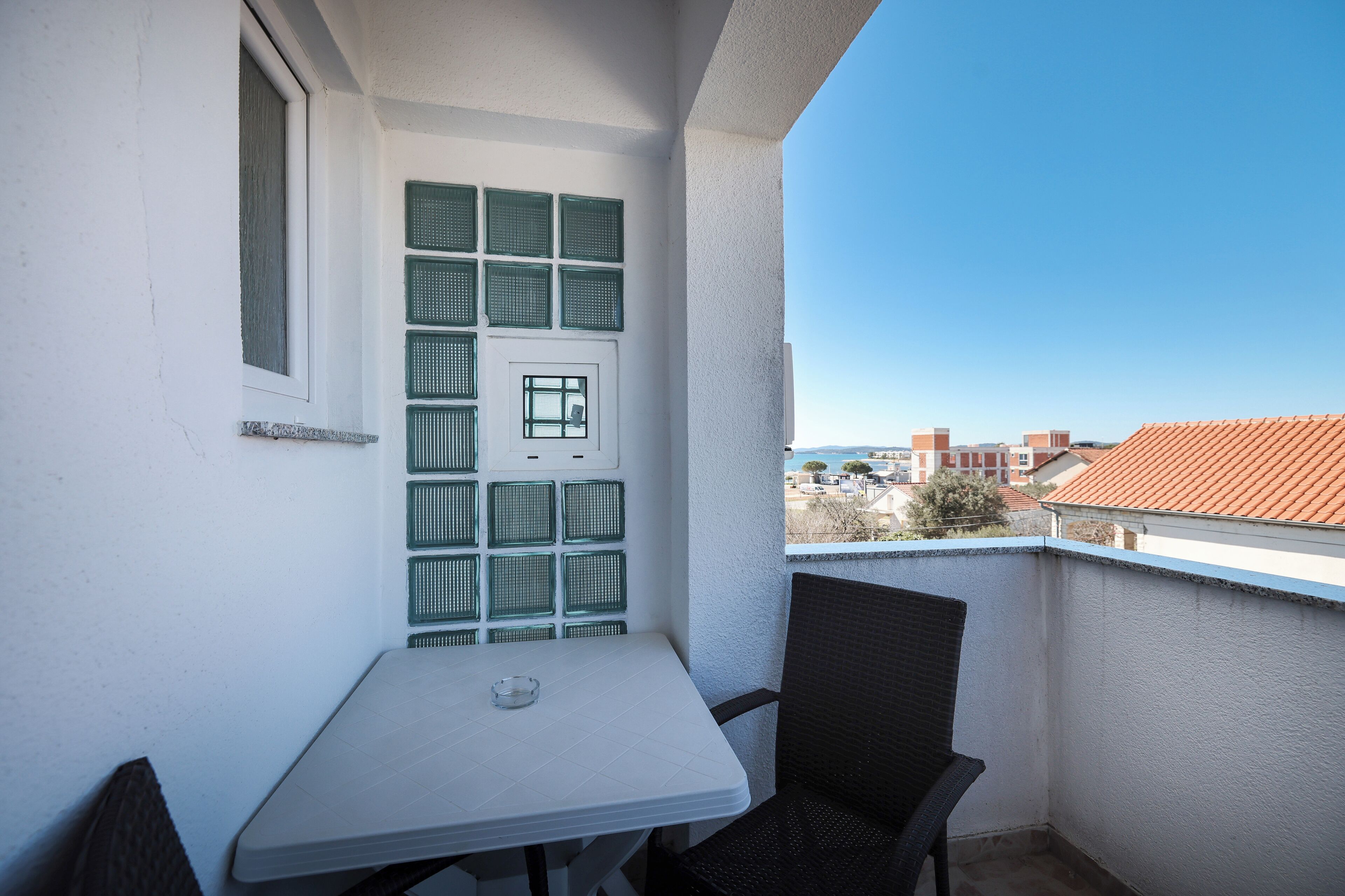 Un balcón con una mesa blanca y dos sillas, una ventana con contraventanas verdes y vistas a edificios y árboles.