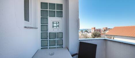 A balcony with a white table and two chairs, a window with green shutters, and a view of buildings and trees.