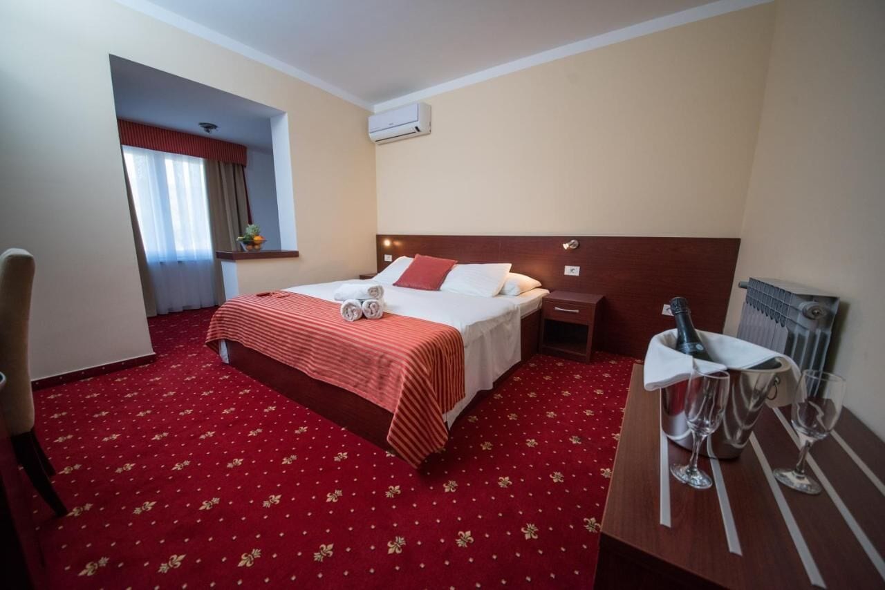 Une chambre d’hôtel avec un lit, une table de chevet sur laquelle se trouvent une bouteille et des verres, une chaise et une moquette rouge.