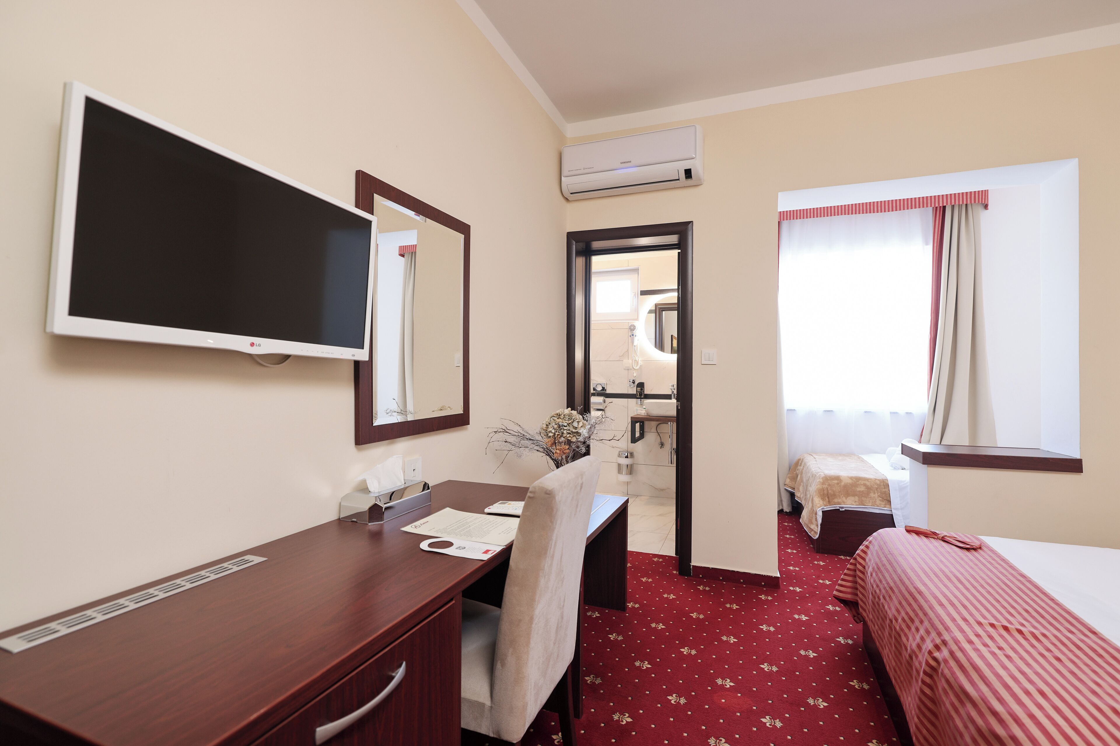 Quarto de hotel com escrivaninha, cama, televisão, espelho, janela com cortinas e ar condicionado.