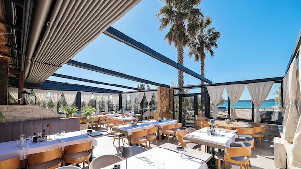 An outdoor dining area with tables and chairs, a palm tree, and a view of the sea. - Villa Anton B&B (Sukošan)