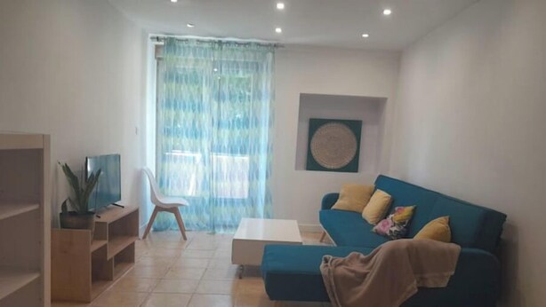 Una sala de estar con un sofá azul, una mesa de centro blanca, un televisor sobre un mueble de madera y un ventanal con cortinas de encaje.