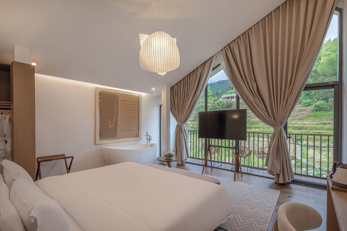 A modern bedroom with a large bed, a flat-screen TV, a balcony with a view of greenery, and a large window with curtains.