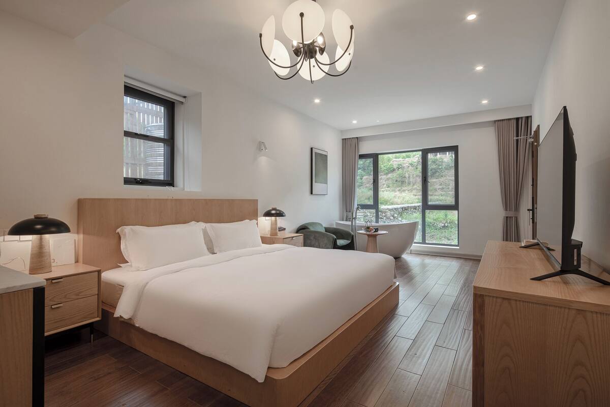 A modern hotel room with a large bed, a desk, a chair, a vanity, and a large window offering a view of greenery.