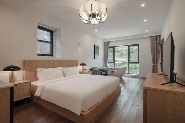 A modern hotel room with a large bed, a desk, a chair, a vanity, and a large window offering a view of greenery.