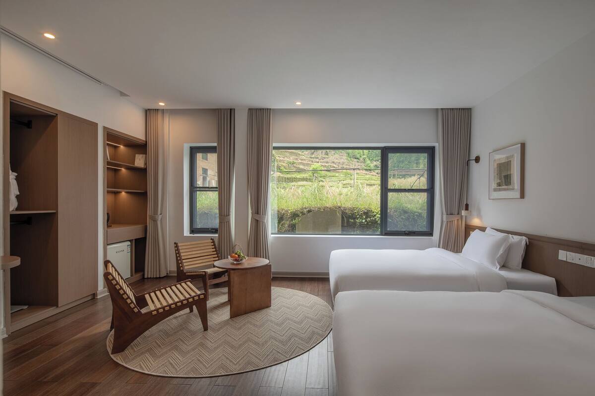 A hotel room with two beds, a wooden chair, a small table, and a window with a view of greenery.