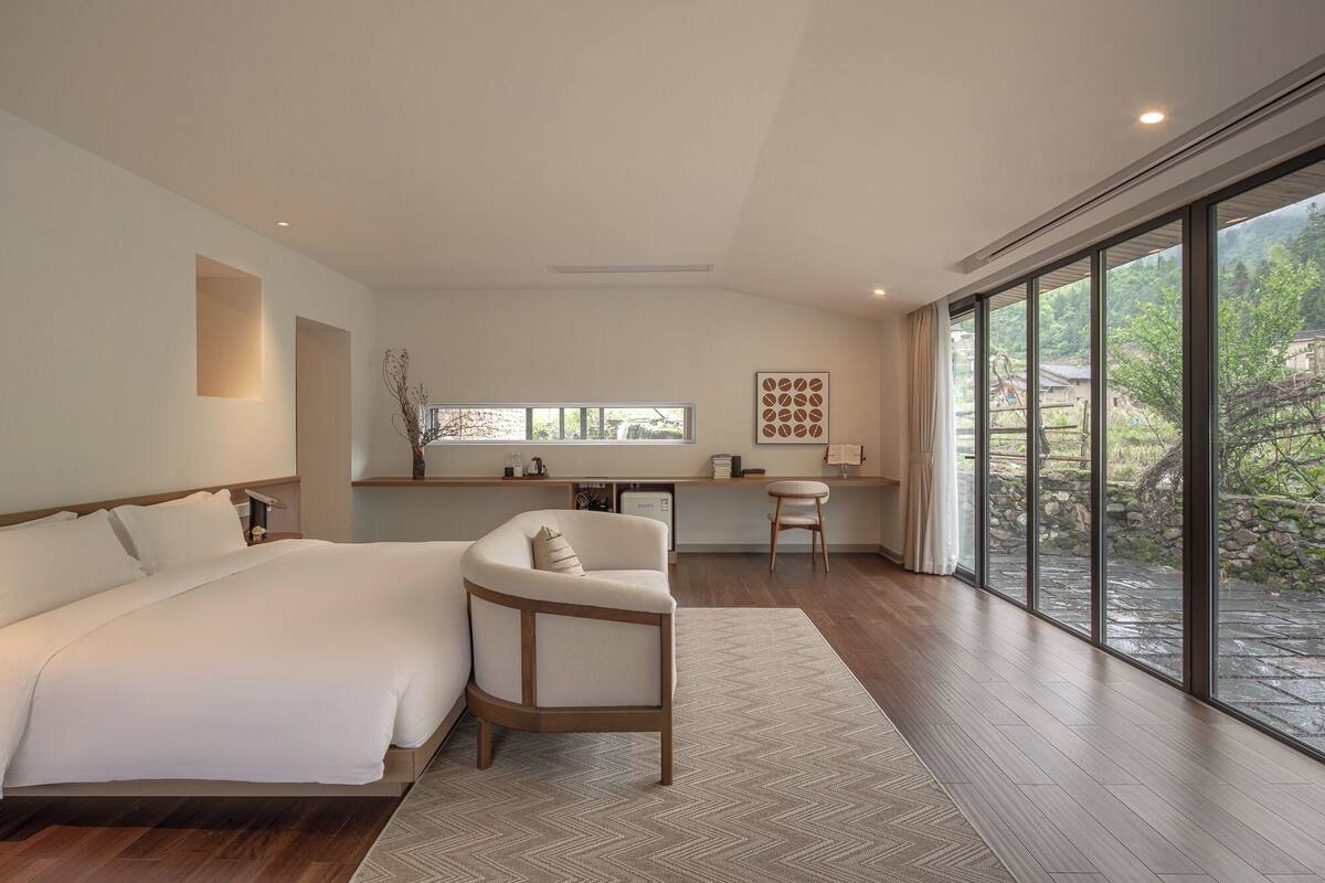 A modern bedroom with a large bed, a desk, and a view of a garden through large windows.