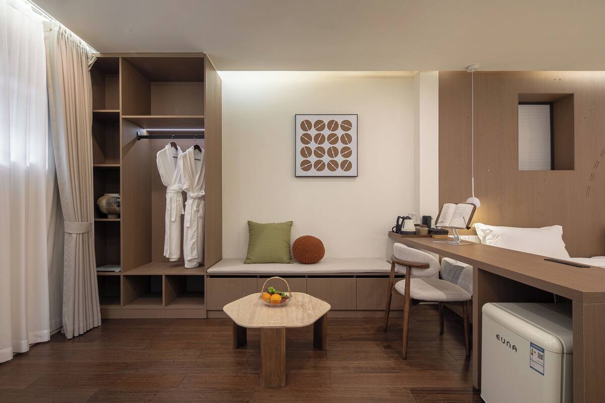A modern hotel room with a wooden desk, a small seating area with a basket of fruit, a green pillow, and a wall-mounted artwork.