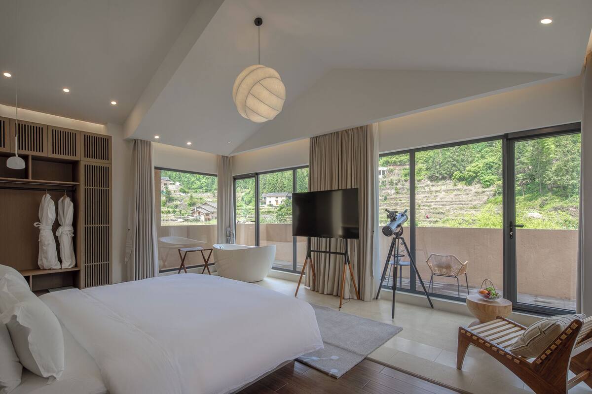 A modern bedroom with a large bed, a sitting area with a TV, and a balcony with a view of greenery.