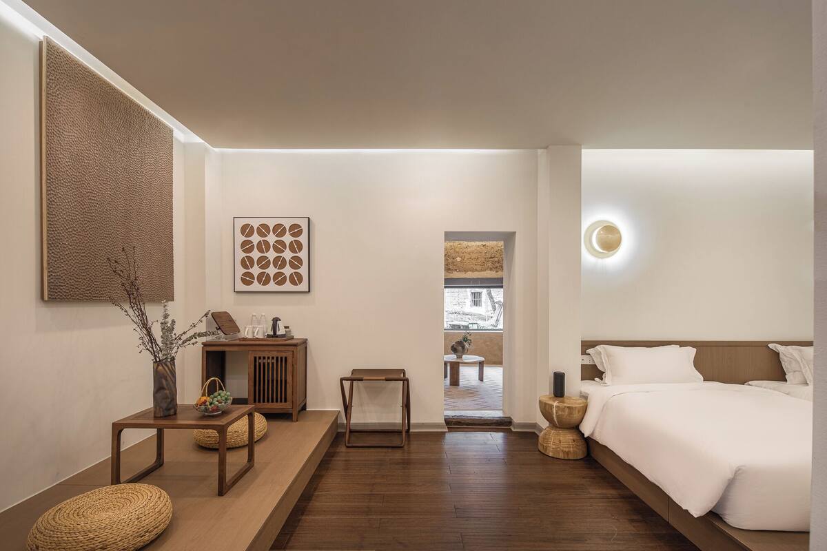 A modern hotel room with a wooden floor, a large bed, a small table with a vase, and a view of the outside through a window.