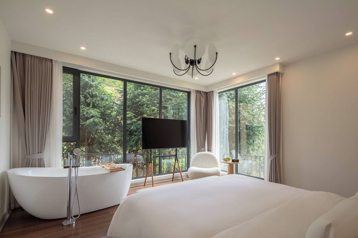 A modern bedroom with a large bed, a freestanding bathtub, a television, and a view of the forest through large windows.
