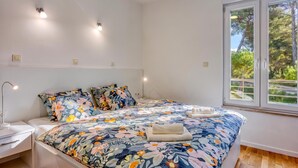 Ein Schlafzimmer mit floralem Bettbezug, Nachttischlampen, einem Fenster mit Blick auf Bäume und einem Holzboden.