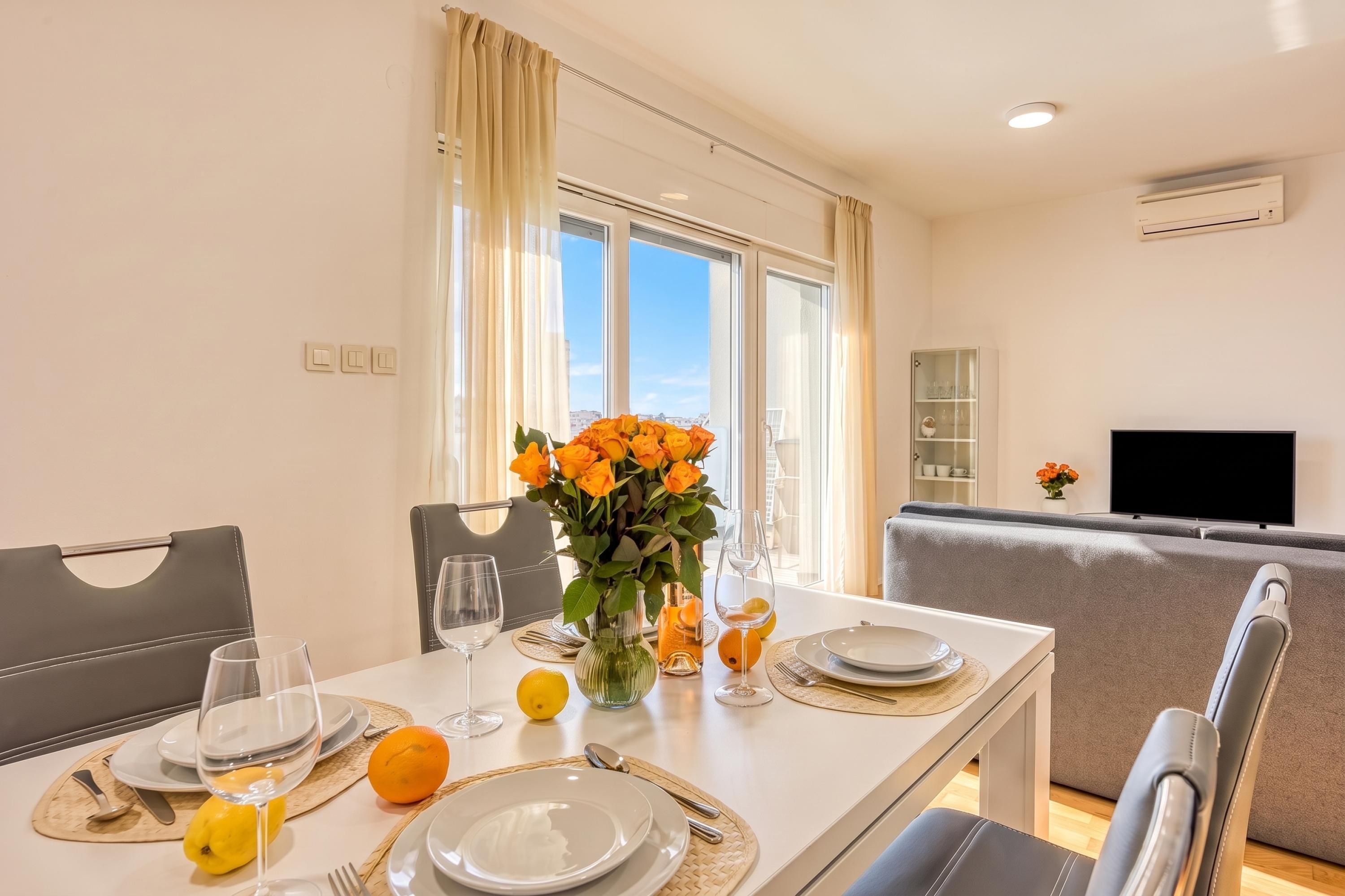 Un coin repas avec une table dressée pour deux personnes, un vase rempli de fleurs et un téléviseur en arrière-plan.