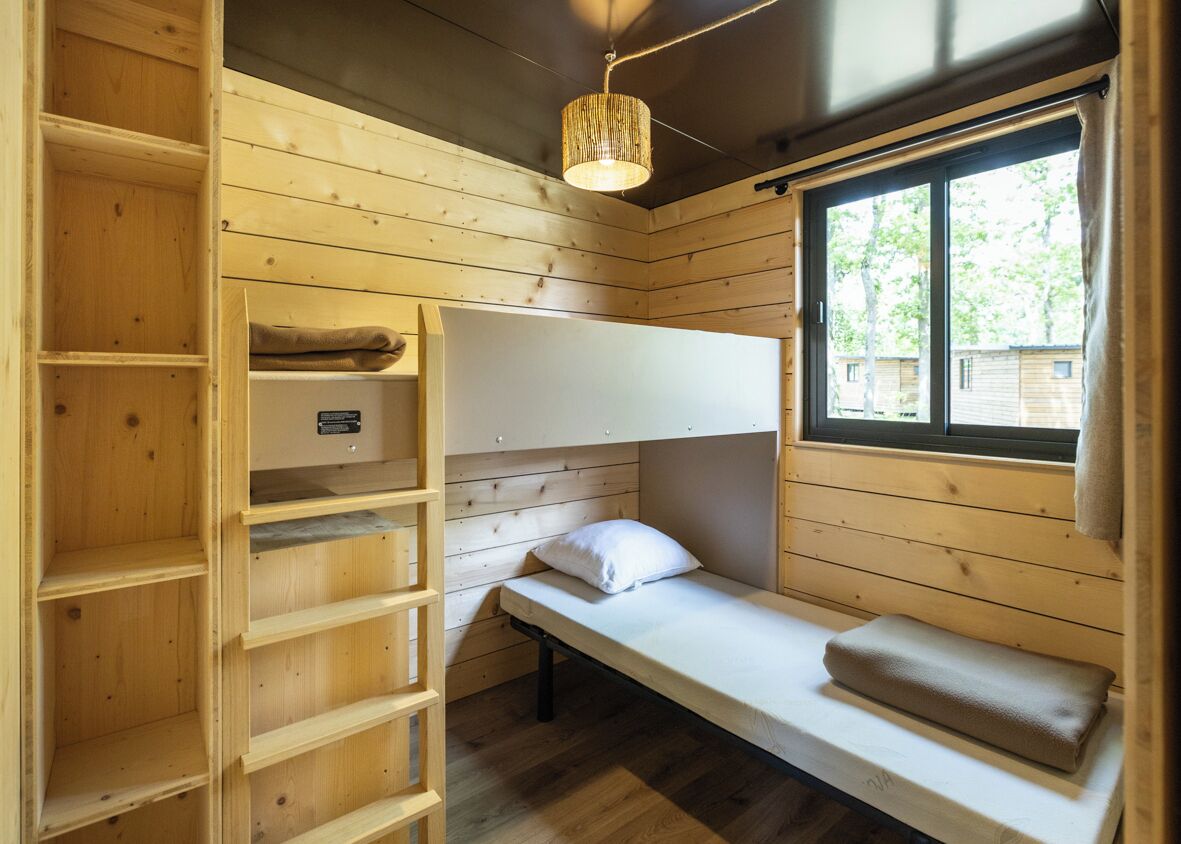 Une cabane aux murs en bois, avec un lit superposé, une étagère et une fenêtre donnant sur les arbres.