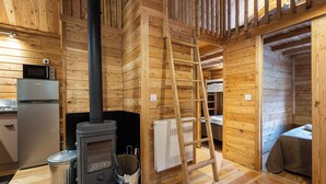 Een houten blokhut van binnen met een houtkachel, een ladder, een klein keukentje en een slaapgedeelte.