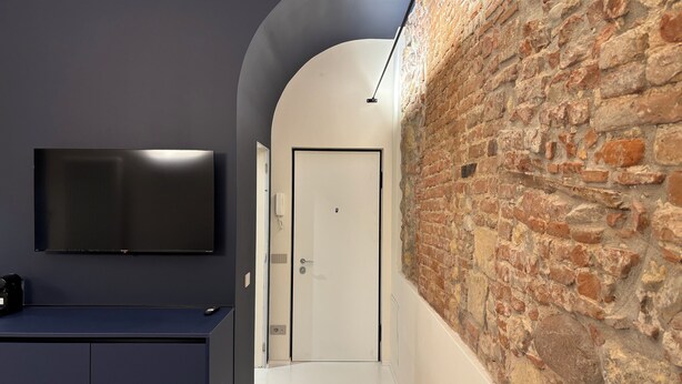 A hallway with a TV mounted on the wall, a door at the end, and a brick wall on the right.