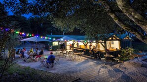 An outdoor dining area with tables and chairs, a covered seating area, and a building with a sign. - Huttopia Oléron les Chênes Verts  (Dolus-d'Oléron)