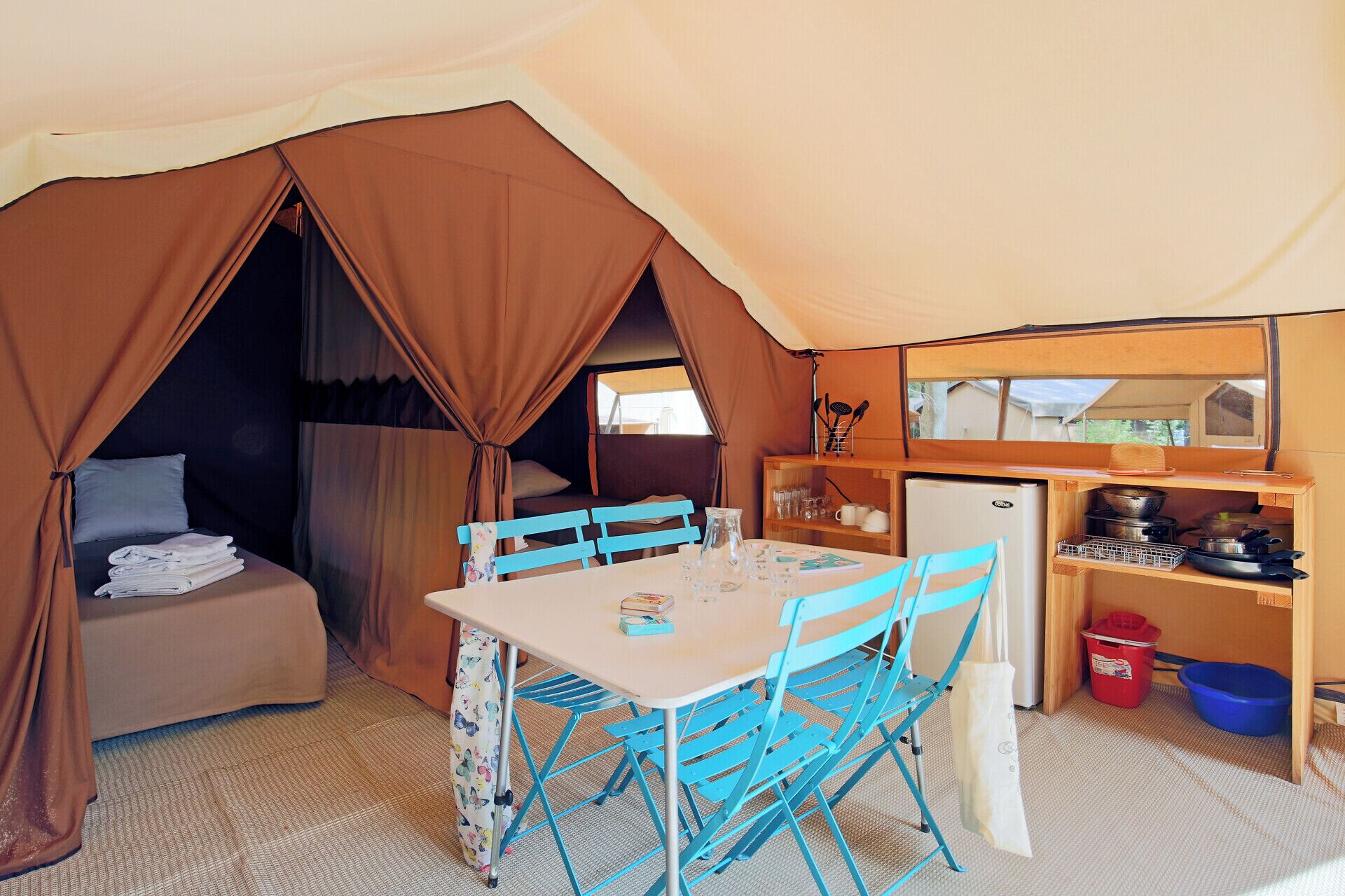 Acomodação em tenda com cama, mesa e cadeiras, pequena cozinha e prateleira para guardar pertences.