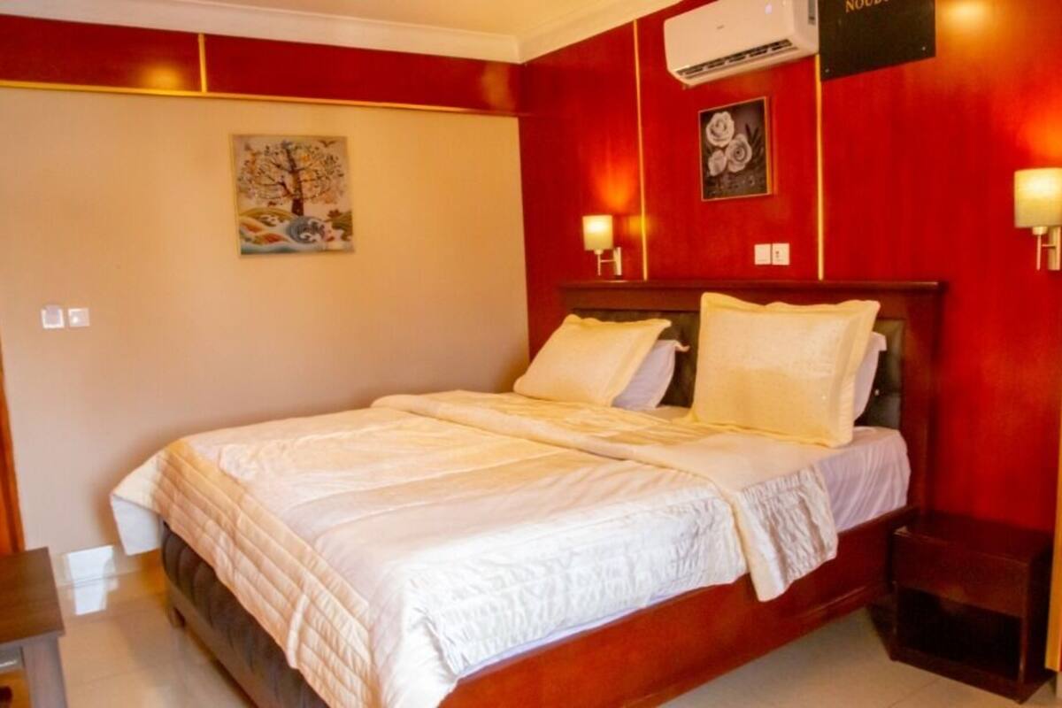 A hotel room with a large bed, a nightstand, a wall-mounted air conditioner, and a painting on the wall.