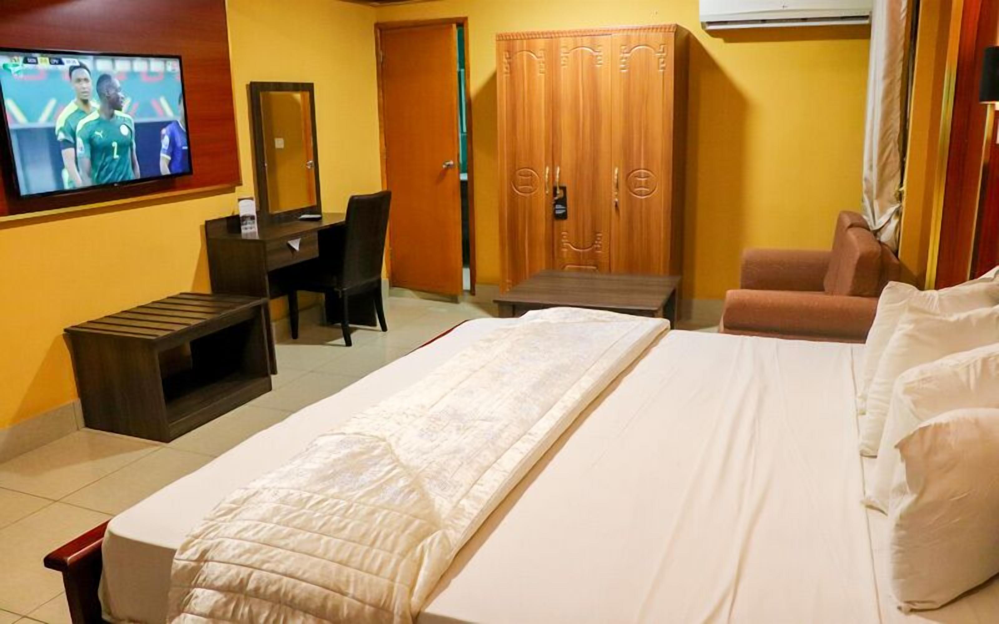 A hotel room with a bed, a desk, a chair, a wardrobe, and a television displaying a sports event.