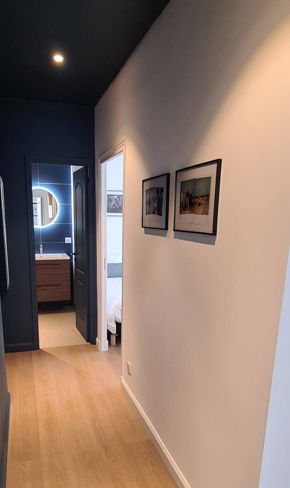 A hallway with a wooden floor, a door leading to a bathroom with a mirror, and two framed pictures on the wall.