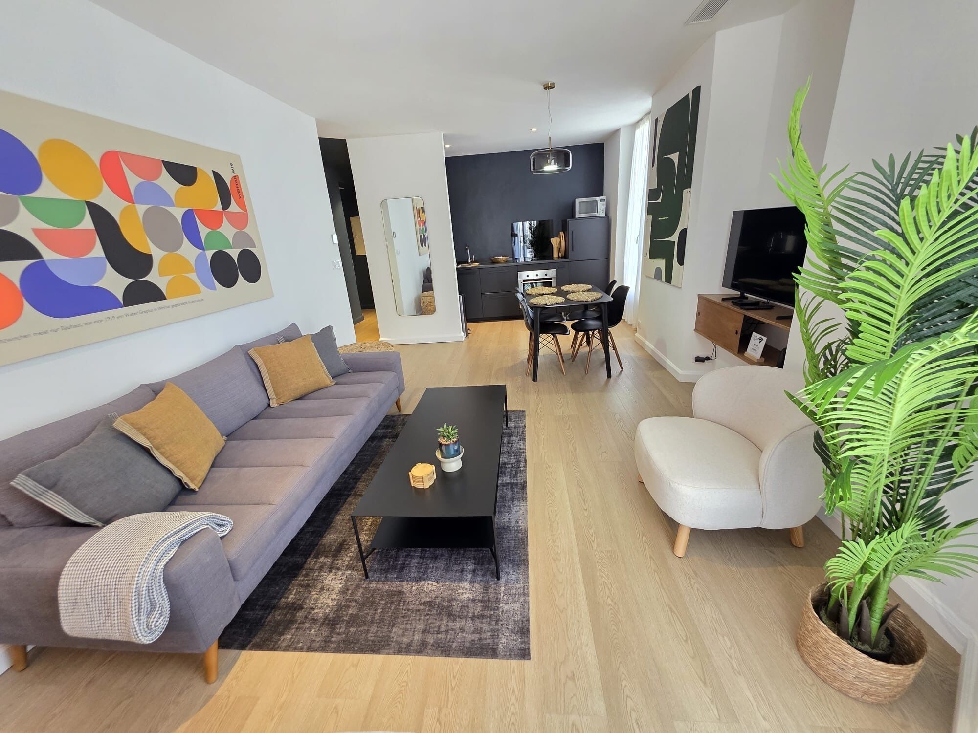 A modern living room with a grey sofa, a black coffee table, and a white armchair. There is a large abstract painting on the wall, a potted plant, and a kitchen area in the background.