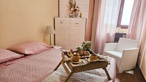 A cozy room with a bed, a wicker chair, a wooden side table with breakfast items, and a cabinet with decorative flowers.