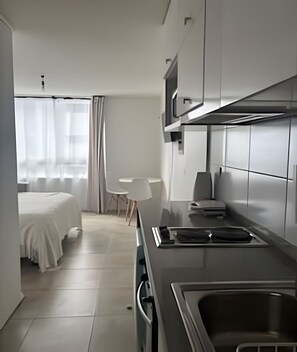 A modern, minimalist hotel room with a bed, a small table, a chair, a kitchenette, and a window with sheer curtains. - Hermoso estudio en Santiago (Santiago)