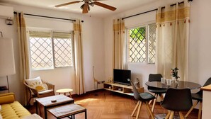 Een woonkamer met een bank, een salontafel, een televisie op een standaard, een eettafel met stoelen en grote ramen met gordijnen.