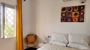 Een slaapkamer met een bed, een stoel, een nachtkastje, een airconditioning en een raam met gordijnen.