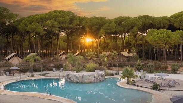 A pool area with a stone feature, lounge chairs, and umbrellas, surrounded by trees and a sunset view. - Huttopia Parque de Doñana  (Hinojos)