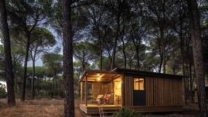 A small cabin with a glass door, surrounded by trees, and a wooden deck. - Huttopia Parque de Doñana  (Hinojos)