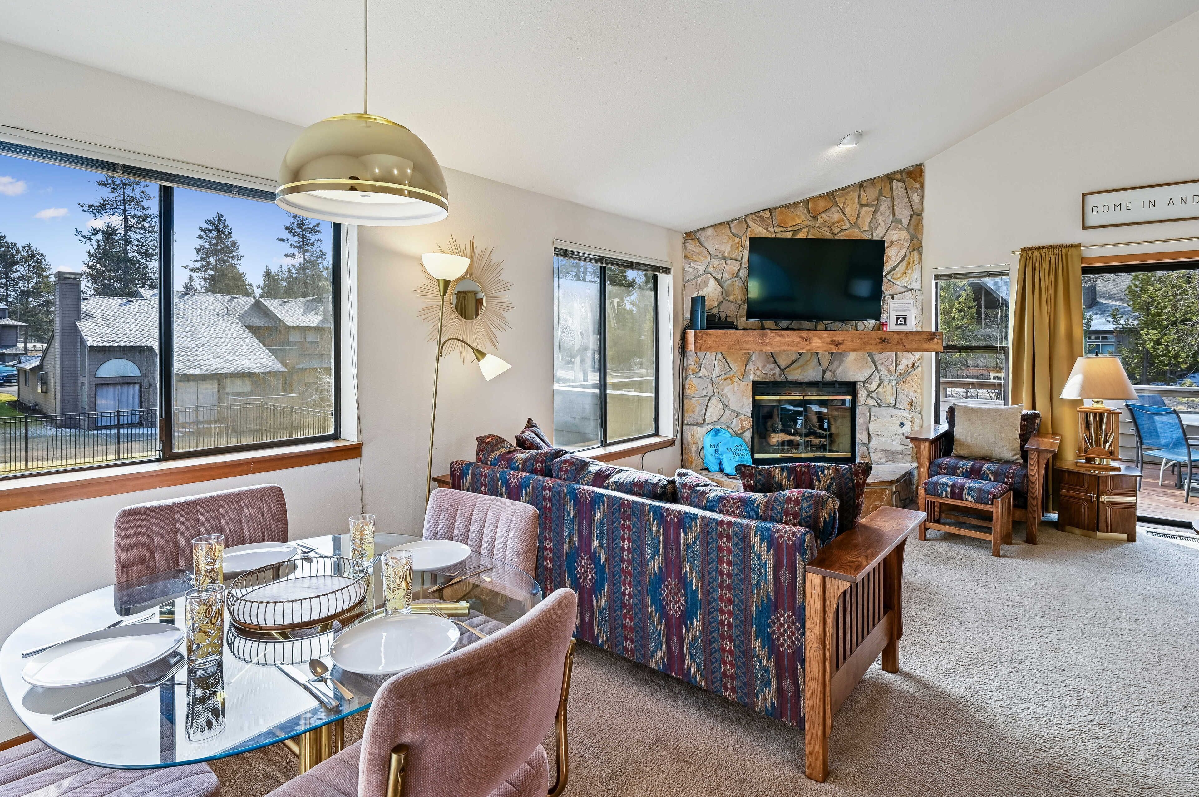 A modern living room with a glass dining table, comfortable seating, a stone fireplace, and a large TV.