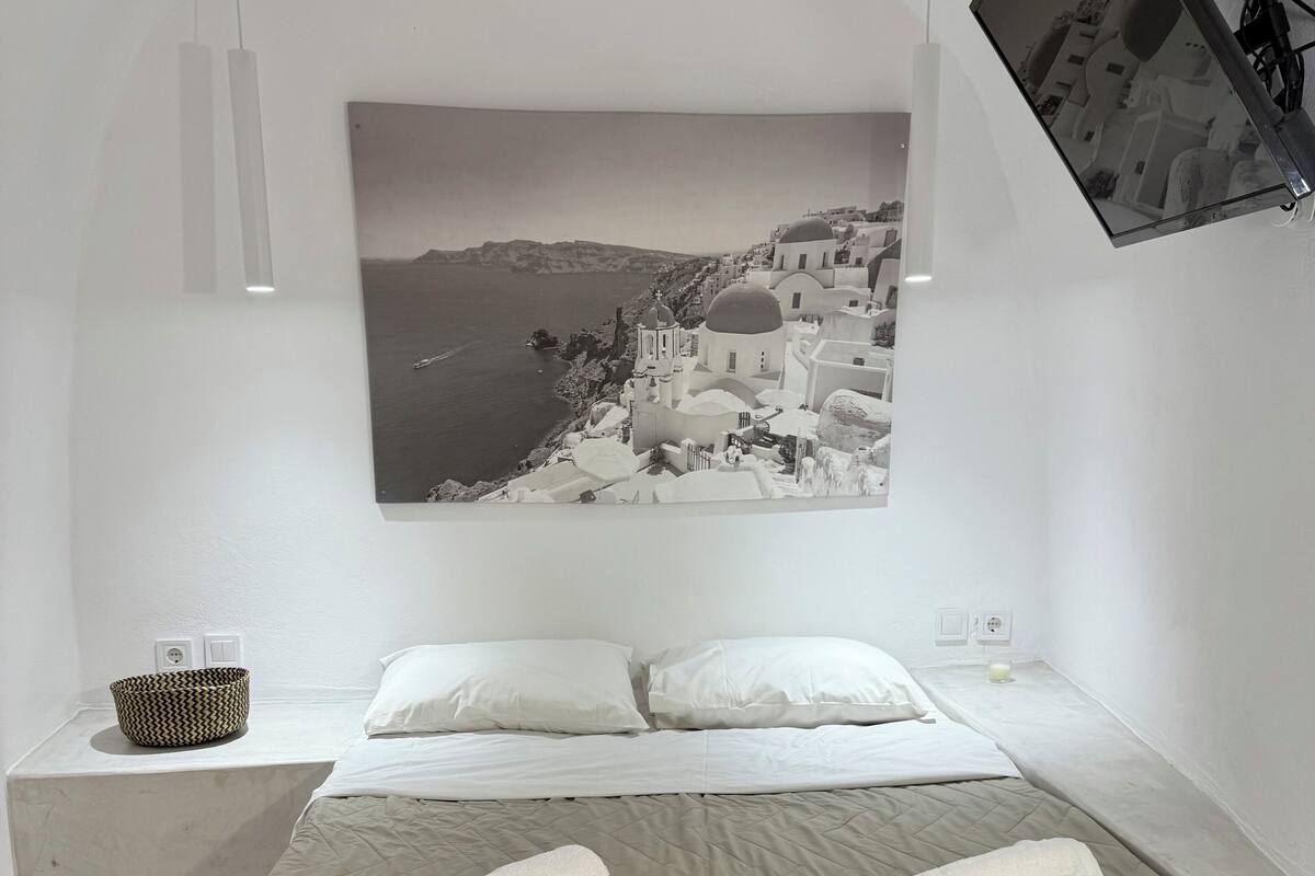 A minimalist hotel room with a bed, two pillows, a bedside table with a basket, and a wall-mounted picture of a coastal town.