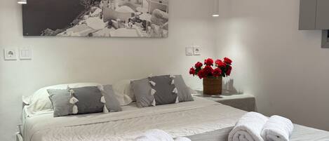 A neatly made bed with white linens and rolled towels, a gray and white patterned bedspread, a wall-mounted painting of a coastal town, and a small vase with red flowers on a bedside table.