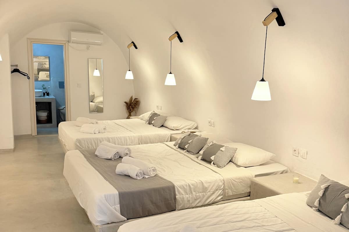 A hotel room with three beds, each with white linens and grey pillows. There are wall-mounted lights and a visible bathroom with a sink and mirror.