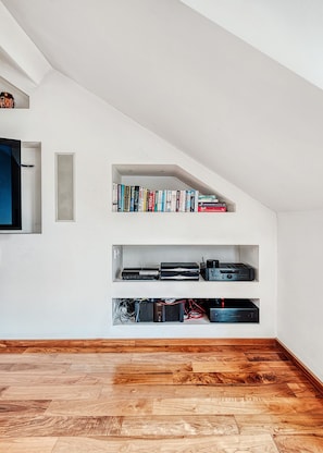 Ein Zimmer mit schrägem Dach, Holzboden und eingebauten Regalen mit Büchern und elektronischen Geräten.