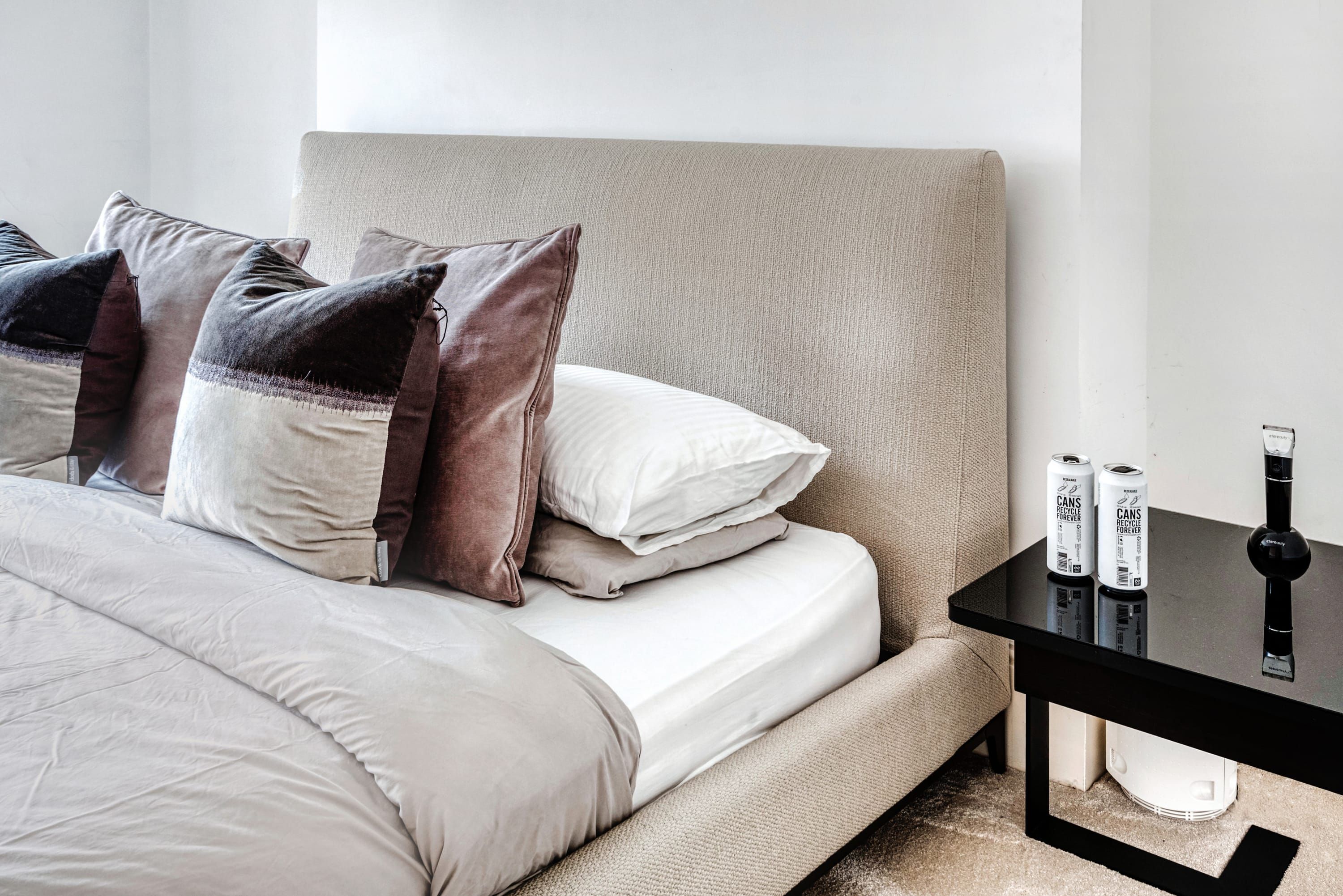 A neatly made bed with a beige headboard, a black side table with two cans of beer, and a white bedside table.